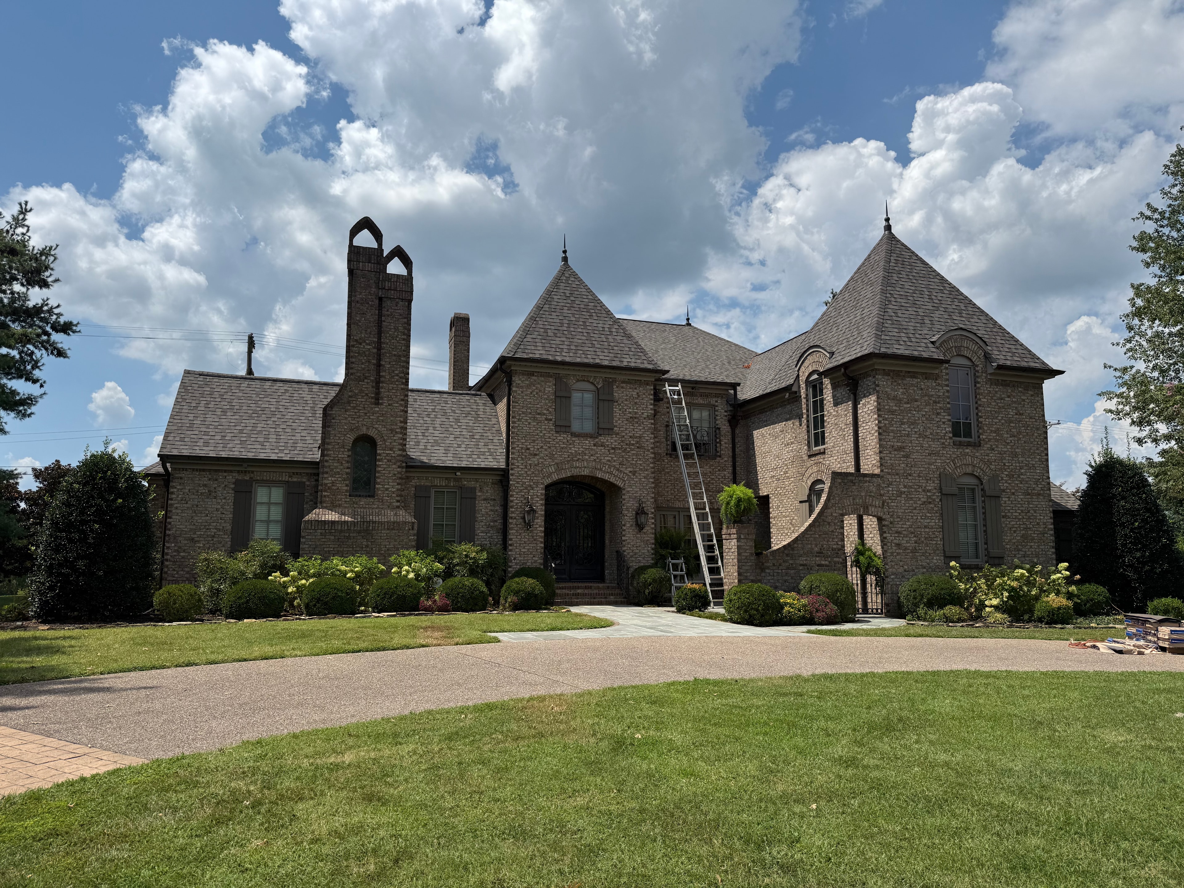 Large brick estate home with completed dark shingle roof and ladder against facade
