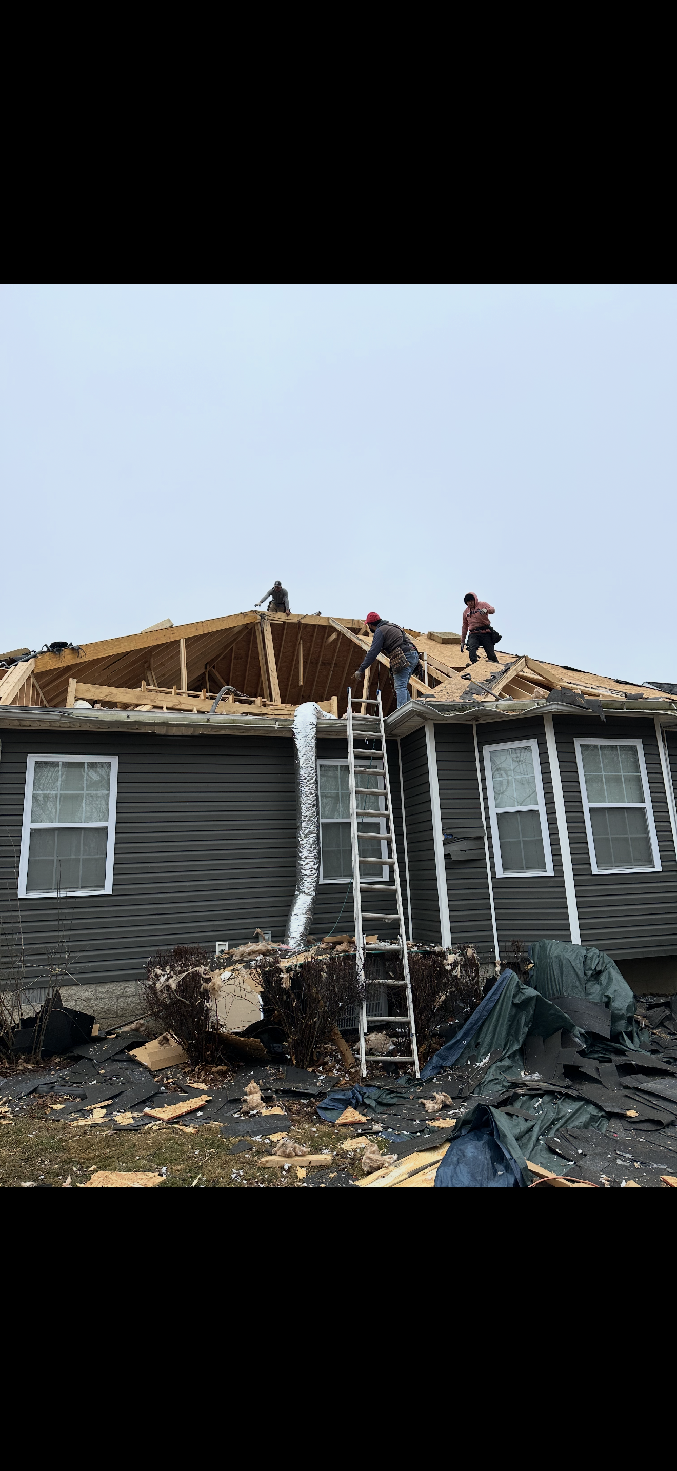 Crew working on storm-damaged gray home with exposed roof decking and debris on ground